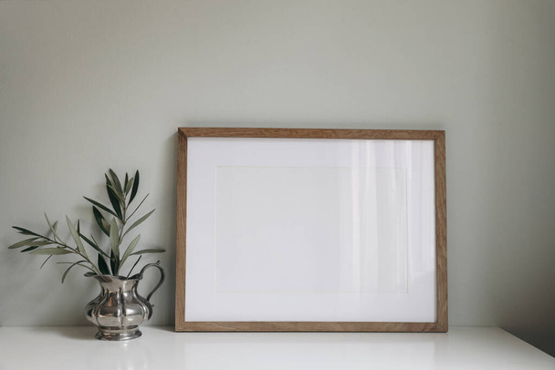 Empty horizontal wooden picture frame mockup. Olive tree branches in silver jug vase. Mint green wall background. Working space, home office. Art, poster display, retro summer interior still life. - 写真・画像