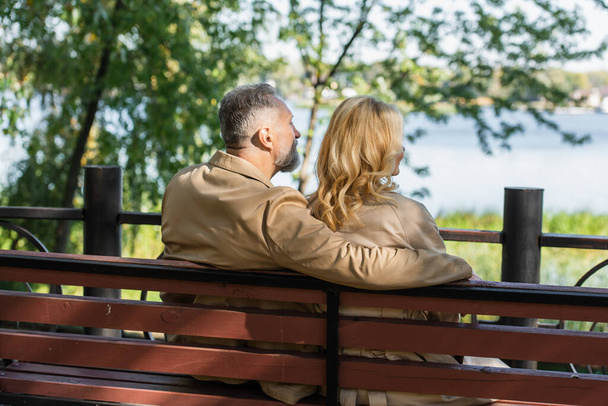 Мужчина средних лет обнимает жену-блондинку, сидя на скамейке в весеннем парке  - Фото, изображение