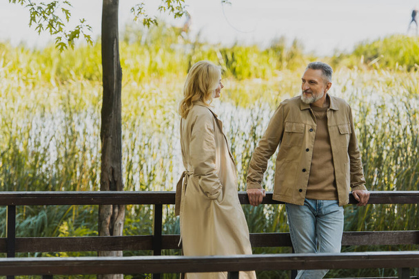 Sonriente hombre maduro mirando rubia esposa en gabardina en puente en parque  - Foto, imagen
