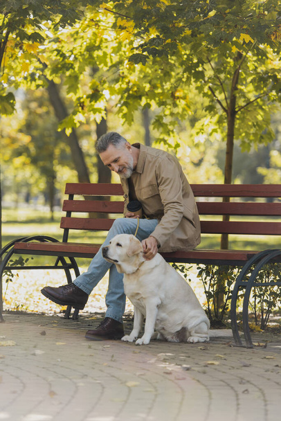 Smiling mature man holding coffee to go and petting labrador in park  - Foto, Imagen