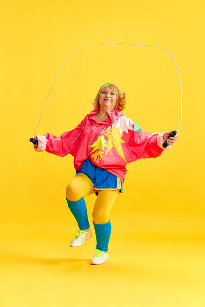 Happy senior sportive woman in colorful uniform training, jumping rope, posing against yellow studio background. Concept of sportive lifestyle, retirement, health care, wellness. Ad - Foto, Bild