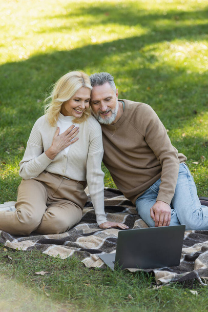 charming middle aged couple watching movie on laptop and sitting on blanket in park  - Foto, immagini