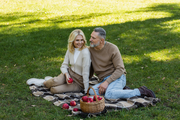 pleased middle aged woman holding ripe apple near bearded husband during picnic in park  - 写真・画像