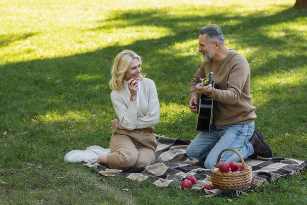 szczęśliwy mężczyzna w średnim wieku z szarą brodą gra na gitarze akustycznej w pobliżu beztroskiej żony z jabłkiem podczas pikniku w parku  - Zdjęcie, obraz