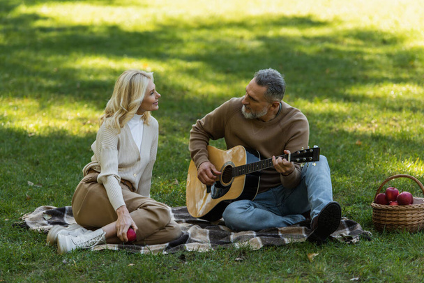 mężczyzna w średnim wieku z szarą brodą gra na gitarze akustycznej w pobliżu uroczej żony podczas pikniku w parku  - Zdjęcie, obraz