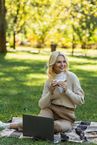 mujer de mediana edad feliz comer sándwich club y sentado en la manta cerca de la computadora portátil durante el picnic en el parque  - Foto, imagen