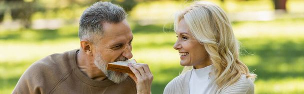 ευτυχισμένη μεσήλικη γυναίκα σίτιση γενειοφόρος σύζυγος με νόστιμο σάντουιτς, πανό  - Φωτογραφία, εικόνα