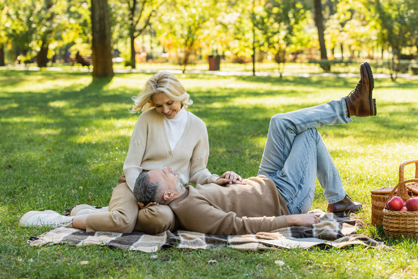 bärtiger Mann mittleren Alters liegt beim Picknick im Park auf dem Schoß seiner glücklichen Frau  - Foto, Bild