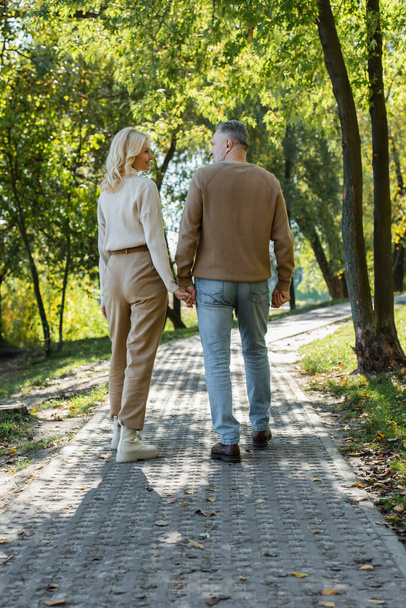 緑の公園を歩いている間に手を携えてスタイリッシュな中年のカップルの完全な長さ  - 写真・画像