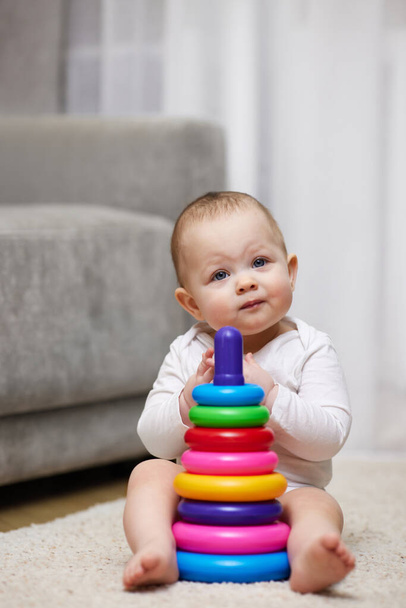 Cute baby girl playing with colorful toy pyramid sitting on carpet at home. Early development for kids. - Фото, изображение