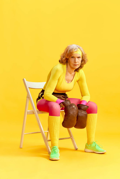 Middle-aged sportive woman in colorful uniform sitting on chair in retro boxing gloves with serious face against yellow background. Concept of sportive lifestyle, retirement, health care, wellness. Ad - Foto, Bild