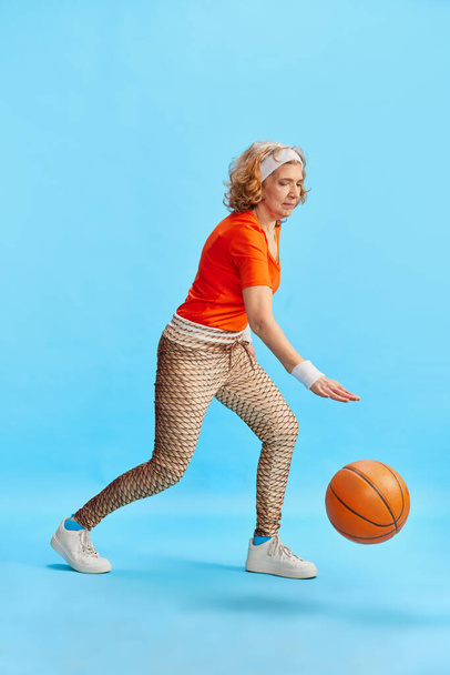 Middle-aged sportive woman in colorful stylish uniform training, dribbling basketball ball against blue studio background. Concept of sportive lifestyle, retirement, health care, wellness. Ad - Zdjęcie, obraz