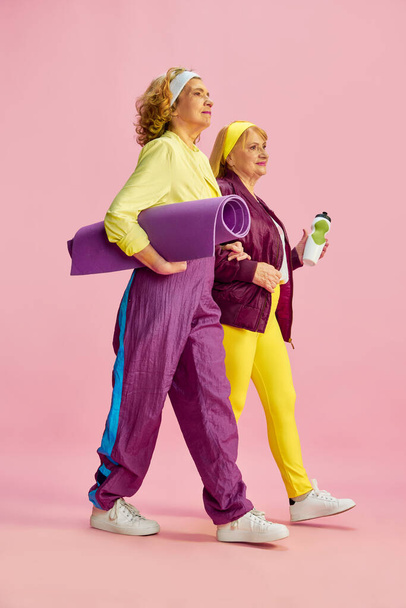 Middle-aged sportive woman in colorful uniform going to gym with fitness equipment, posing against pink studio background. Concept of sportive lifestyle, retirement, health care, wellness. Ad - Zdjęcie, obraz