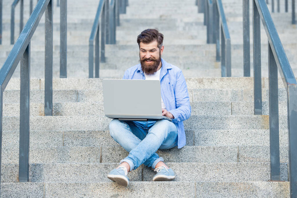 happy guy has online video communication outdoor. guy has online video communication outside. guy has online video communication sitting on stairs. guy has online video communication on laptop. - Photo, image