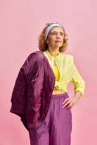 Beautiful middle-aged, sportive woman in colorful uniform posing against pink studio background. Fitness routine. Concept of sportive lifestyle, retirement, health care, wellness. Ad - 写真・画像