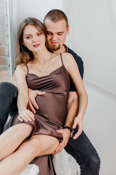 Een man en een zwangere vrouw zitten op een vensterbank en knuffelen elkaar. - Foto, afbeelding