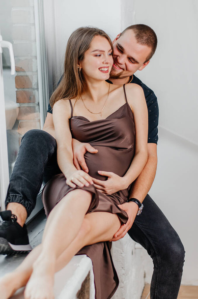 Ein Mann und eine schwangere Frau sitzen auf einer Fensterbank und umarmen sich - Foto, Bild