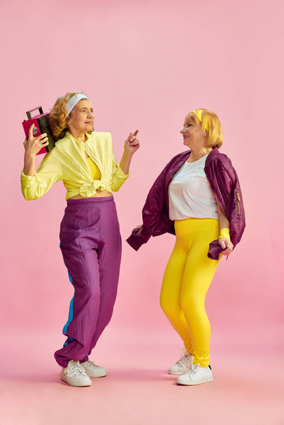 Two middle-aged, elderly, sportive woman in colorful uniform listening to radio and dancing, posing against pink studio background. Concept of sportive lifestyle, retirement, health care, wellness. Ad - Fotografie, Obrázek