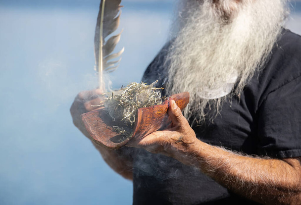 人間の手は青い背景を背景にオーストラリアの植物の枝と羽を持つ木製の料理を保持し、オーストラリアの先住民コミュニティイベントで煙の儀式の儀式 - 写真・画像