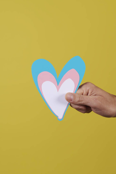 close-up da mão de um jovem caucasiano segurando um coração com as cores da bandeira transgênero em um fundo amarelo - Foto, Imagem