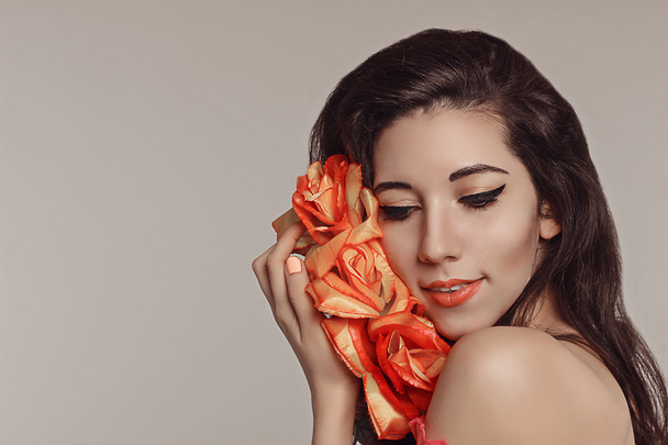 Весняна модель студійної зйомки. Портрет молодої красивої жінки брюнетки з квітами червоних троянд на білому тлі. Модний макіяж. Чорні стріли і чуттєві губи. Чиста шкіра. Ніжність
 - Фото, зображення