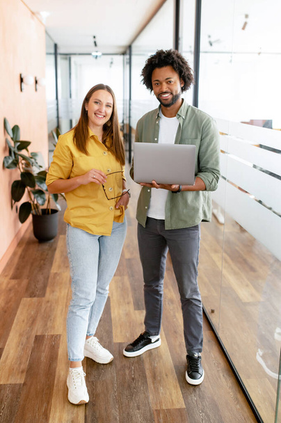 Two coworkers man and woman is using laptop, standing at the office. They holding laptop, looking at camera and smiling, full lengths, friendly office atmosphere and teamwork concept - Foto, Imagen