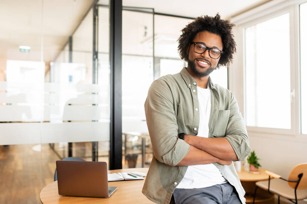 Ambitious highly-skilled handsome curly african-american male employee in glasses and casual wear leaned on office desk and looking at the camera with smile, businessman stands with arms crossed - Φωτογραφία, εικόνα