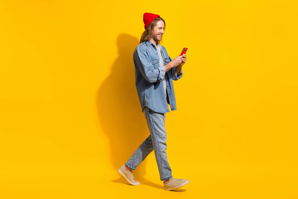 Full size photo of positive man wear denim jacket jeans trousers hold smartphone walk empty space isolated on yellow color background. - Foto, imagen