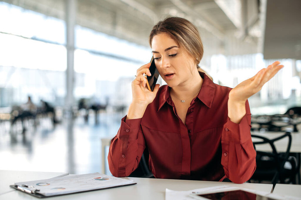 Busy confident caucasian woman, business lady, sits at workplace in the office, has a working conversation by smart phone with a colleague or client, discusses work issues, gesticulates with her hand - Photo, Image