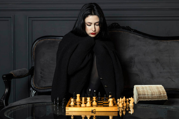 Retrato de una chica hermosa y elegante en negro con lápiz labial rojo mate brillante, se sienta detrás de un tablero de ajedrez de madera, se cubrió con un suéter, mirando cuidadosamente las piezas. - Foto, Imagen