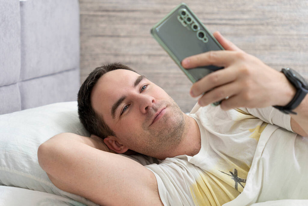Smiling handsome man lying on bed and looking at his smartphone at the morning time. Reading news and chatting by using phone concept - Foto, Imagen