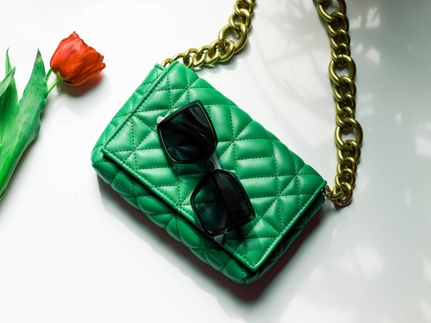 Stylish women's trendy green quilted bag with a large gold chain, a red tulip in it and sunglasses lie on a white table with shadows from the window, flat lay close-up. Fashionable accessories concept. - Foto, Imagem
