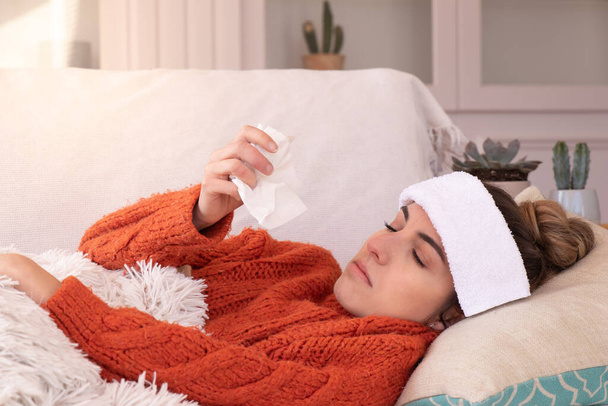Ill female with influenza and fever blowing red running nose while lying on comfortable sofa in light living room - Фото, изображение