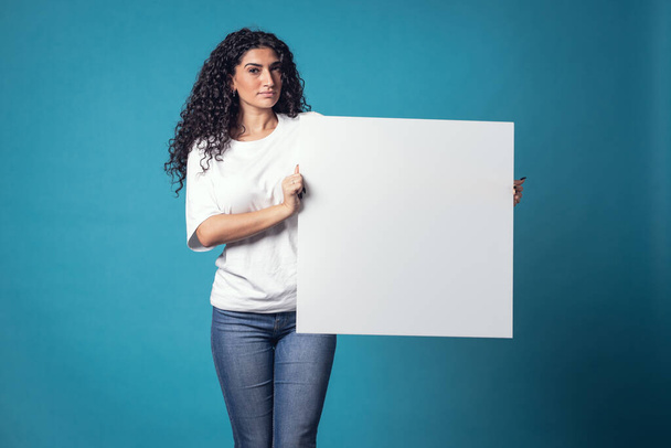 Mooie krullende vrouw houden blanco billboard geïsoleerd op blauwe achtergrond - Foto, afbeelding