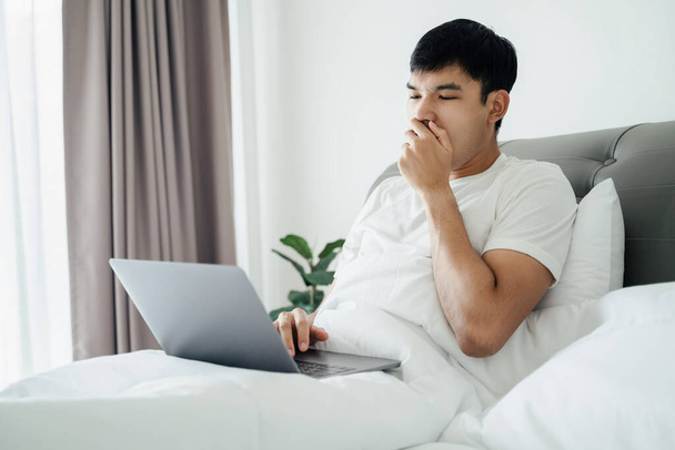 Сонный человек зевает на кровати с помощью ноутбука. - Фото, изображение
