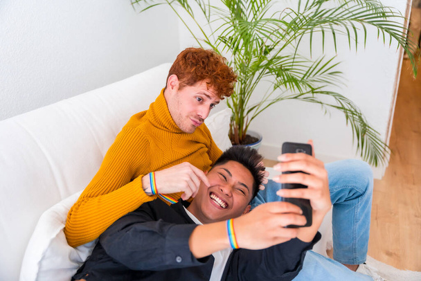 Beautiful gay couple being romantic indoors at home on the sofa, gay couple taking selfie, lgbt concept - Φωτογραφία, εικόνα