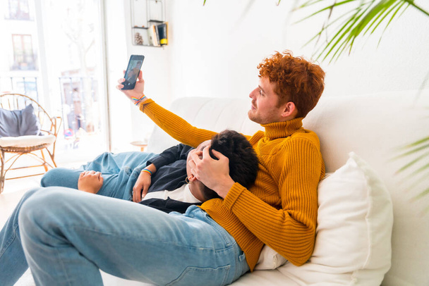 Beautiful gay couple being romantic indoors at home on the sofa, gay couple taking selfie, lgbt concept - Фото, изображение