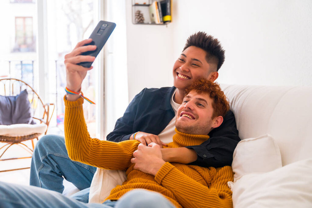Beautiful gay couple being romantic indoors at home on the sofa, taking a selfie, lgbt concept - Fotó, kép