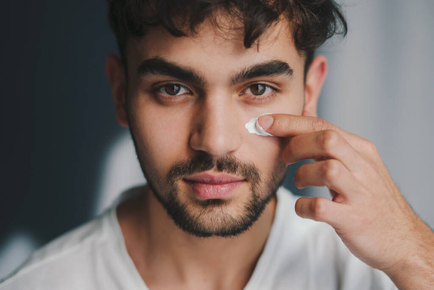 Close up portrait of caucasian man applying moisturizing cream on his face isolated on gray background. People lifestyle. Beauty treatment. Facial beauty. Male - Fotoğraf, Görsel
