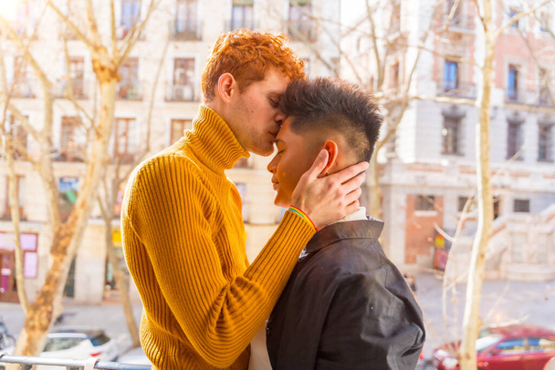 Beautiful gay couple being romantic at home on the sofa, home balcony kissing each other on the forehead, lgbt concept - 写真・画像