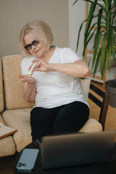 Πορτρέτο ηλικιωμένης γυναίκας που ενυδατώνει το δέρμα των χεριών της καθισμένη σε καναπέ στο σπίτι. Δέρμα ομορφιάς. - Φωτογραφία, εικόνα