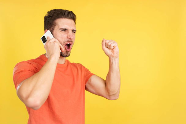 Happy caucasian man celebrating while taking to the mobile in studio with yellow background - Foto, afbeelding