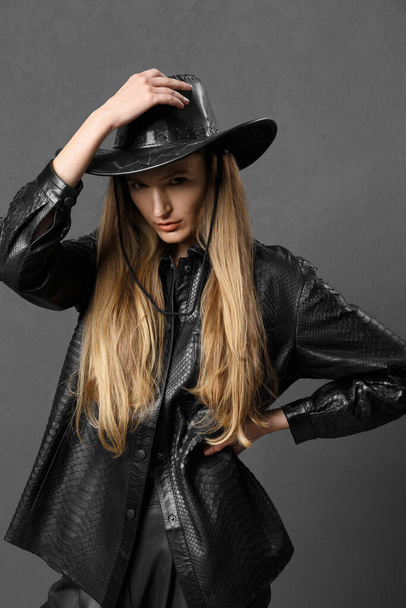 Pretty young sexy fashionable sensual girl posing beautifully on a dark background in a black leather shirt and a cowboy hat, clothing advertisement, a woman in a black leather hat. - Photo, Image