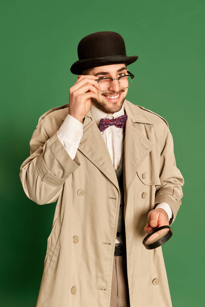 Portrait of young smiling man in stylish trench coat and hat posing with magnifying glass against green studio background. Concept of emotions, profession of detective, secrets, fun, fashion. Ad - Foto, afbeelding
