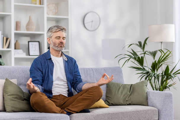Adulto hombre maduro solo en casa relajante sentado en posición de loto en el sofá, persona de pelo gris relajante meditando en el interior de la sala de estar en ropa casual. - Foto, imagen