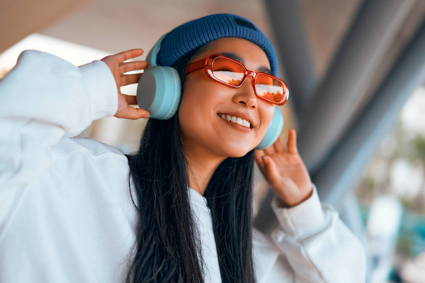 Стильная азиатская молодая женщина в шляпе слушает музыку в наушниках на улице. - Фото, изображение