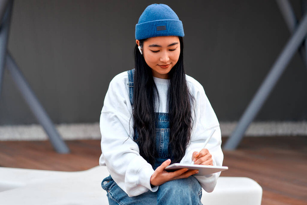 Cute Asian young woman in a hat with a tablet on the street. Drawing on a graphics tablet. Freelancing and earning online. - Photo, Image