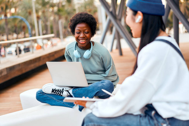 Mujeres jóvenes afroamericanas y asiáticas sentadas con piernas cruzadas al aire libre con computadora portátil y tableta. Freelancing y ganar en línea. - Foto, imagen