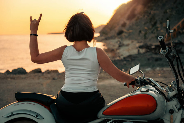 Cool woman posing on motorcycle and show a goat gesture. Back view. Mountain and sunset sky at the background. The concept of Motorcyclist Day. - Photo, Image