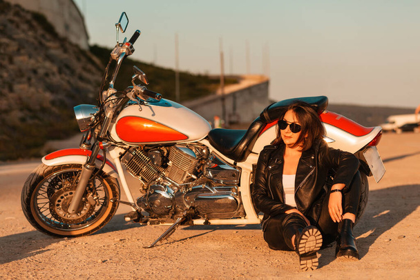 Cool confident woman in a leather motorcycle outfit sitting on the ground near a motorcycle. Sunlight. The concept of the Motorcyclist Day. - Fotoğraf, Görsel